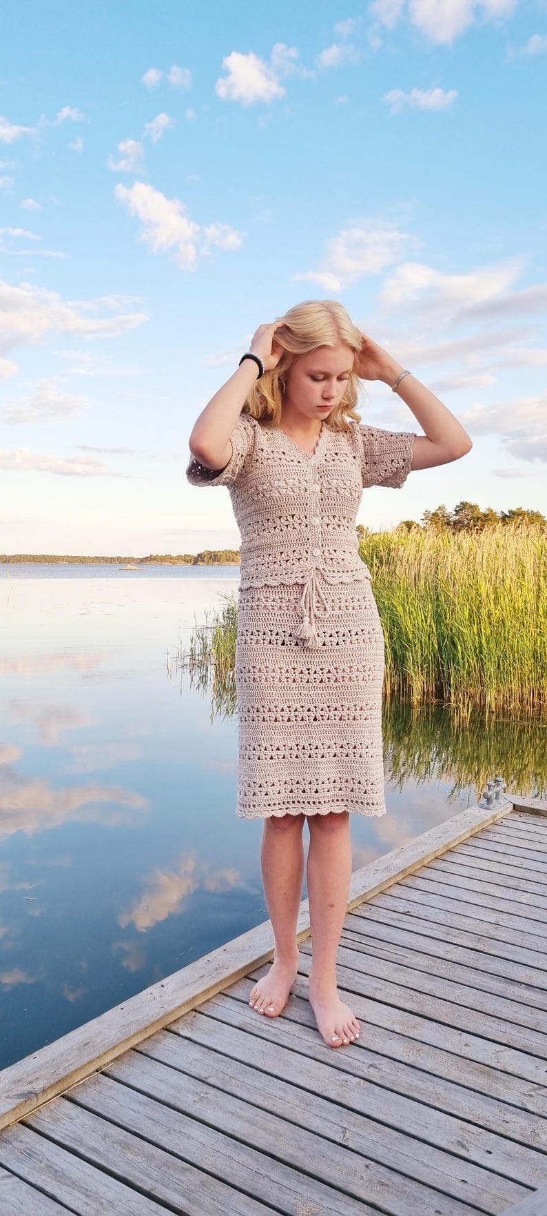 May include: A beige crochet dress with a matching short-sleeved top and knee-length skirt. The dress features a button-down front and a drawstring waist. The person is standing barefoot on a wooden dock near a body of water.