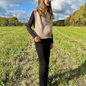 May include: A woman wearing a beige knitted vest with a cable knit pattern over a black long-sleeved shirt and black trousers. She is standing in a field with green grass and a blue sky with white clouds.