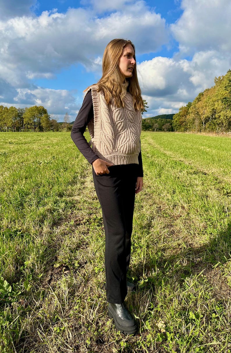 May include: A woman wearing a beige knitted vest with a cable knit pattern over a black long-sleeved shirt and black trousers. She is standing in a field with green grass and a blue sky with white clouds.