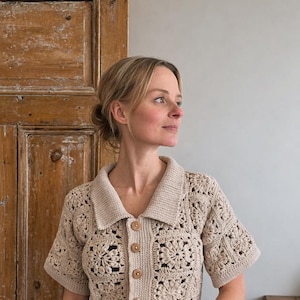 May include: A beige crochet collared shirt with short sleeves and wooden buttons. The shirt features a floral pattern and is buttoned up. The background includes a wooden door and a light grey wall.