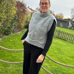 May include: A grey knitted sleeveless turtleneck sweater with a leaf pattern. The sweater is worn over a black long-sleeved shirt and black trousers.