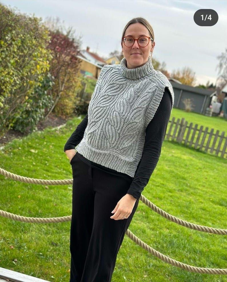 May include: A grey knitted sleeveless turtleneck sweater with a leaf pattern. The sweater is worn over a black long-sleeved shirt and black trousers.