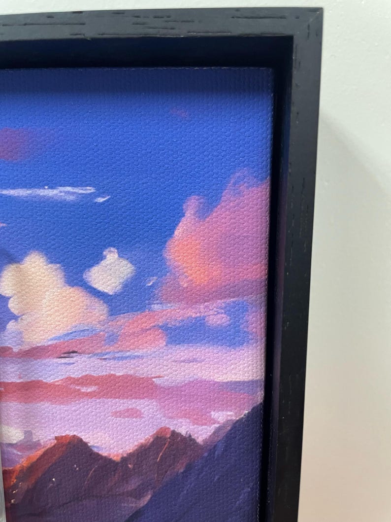 May include: A framed canvas art piece depicting a sunset scene. The artwork features a vibrant sky with shades of blue, pink, and purple, with clouds and mountain silhouettes. The frame is black.