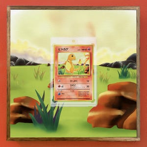 May include: A framed artwork featuring a Pokémon trading card. The card, in a protective sleeve, displays a Charmander illustration with Japanese text. The background depicts a grassy field, rocks, and a sunset sky, all within a wooden frame.