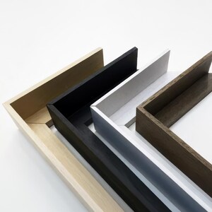 May include: Four different picture frames, each with a different color and wood grain. The frames are stacked on top of each other, with the lightest frame on the bottom and the darkest frame on top. The frames are all made of wood and have a simple, modern design.