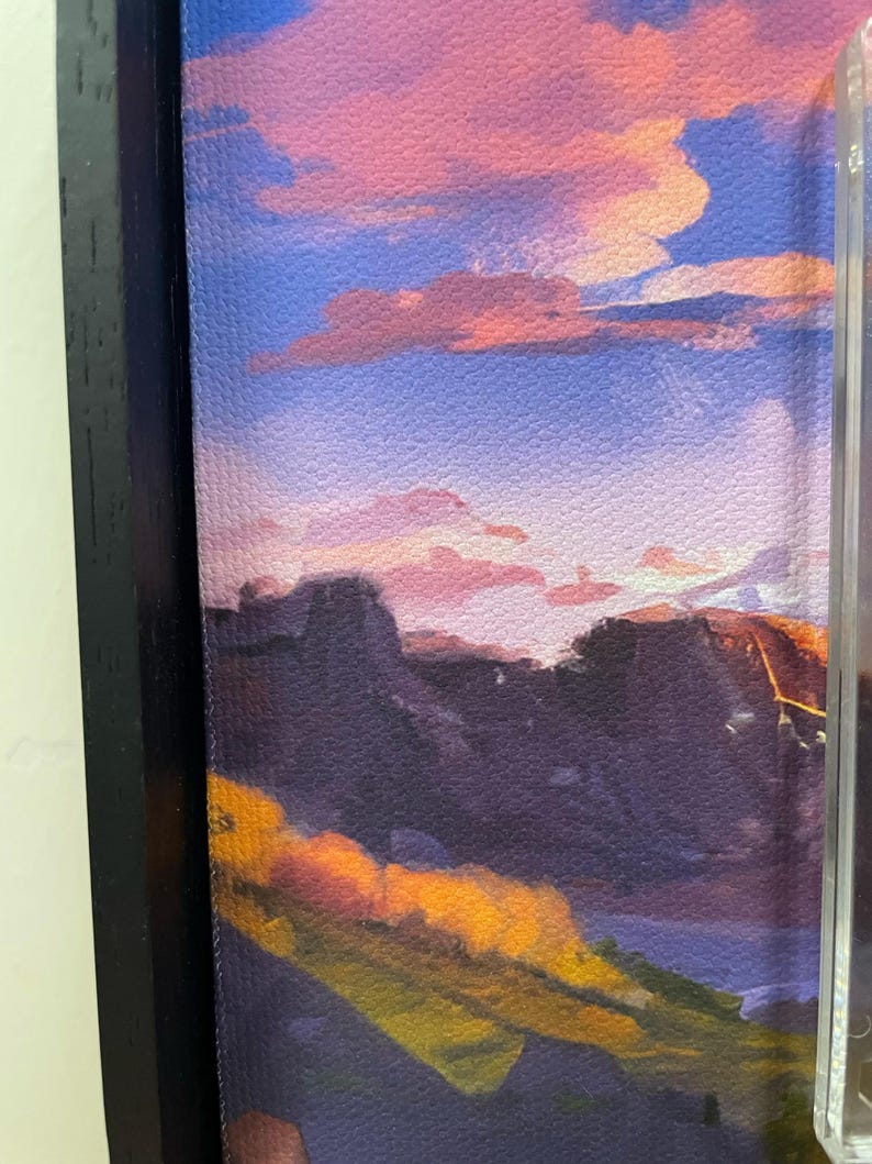 May include: A framed canvas print featuring a landscape scene with a sunset. The artwork displays a vibrant sky with pink and orange clouds above dark purple mountains and yellow and green foliage. The frame is black.