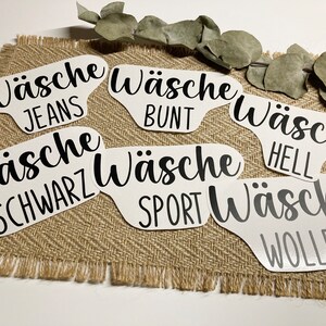Könnte beinhalten: Weiße Wäscheetiketten mit schwarzem Text, darunter "JEANS", "BUNT", "SCHWARZ", "SPORT", "HELL" und "WOLLE". Die Etiketten sind auf einer Jutefläche mit einem Eukalyptuszweig angeordnet.