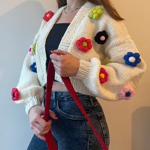 May include: White knitted cardigan with colourful crocheted flower embellishments. The cardigan has a V-neck and long sleeves.