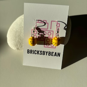 Op de afbeelding: Een paar gele Lego minifiguur hoofd oorbellen met zilveren haakjes. De oorbellen worden getoond op een witte kaart met de tekst "BRICKSBYBEAN".