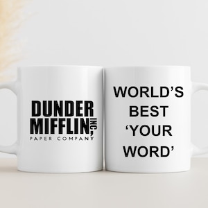 以下が含まれることがあります： 黒い文字が書かれた白いセラミック製のマグカップが2つ。左側のマグカップには「Dunder Mifflin, Inc. Paper Company」と書いてあります。右側のマグカップには「World's Best 'Your Word'」と書いてあります。