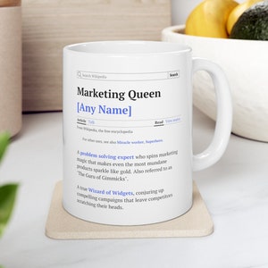 Könnte beinhalten: Eine weiße Keramiktasse mit einem schwarz-weißen Design. Die Tasse zeigt eine Definition von "Marketing Queen" mit dem Text "[Any Name]" anstelle des Namens. Die Definition lautet: "Ein Problemlöser, der Marketing-Magie spinnt, die selbst die banalsten Produkte wie Gold glänzen lässt. Auch bekannt als "Der Guru der Gimmicks". Ein echter Zauberer der Widgets, der überzeugende Kampagnen kreiert, die die Konkurrenz ratlos zurücklassen."