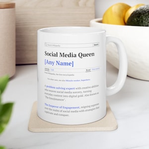 May include: White ceramic mug with a black and white design that reads "Social Media Queen [Any Name]" and a definition of the term. The definition describes a "problem solving expert with creative abilities who weaves social media sorcery, turning everyday content into digital gold. Also known as The Emojimancer." The definition continues, "The Emperor of Engagement, reigning supreme over the realm of social media with strategies that captivate and conquer."