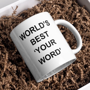 May include: White ceramic mug with black text that reads "WORLD'S BEST 'YOUR WORD'". The mug is sitting in brown packing peanuts.