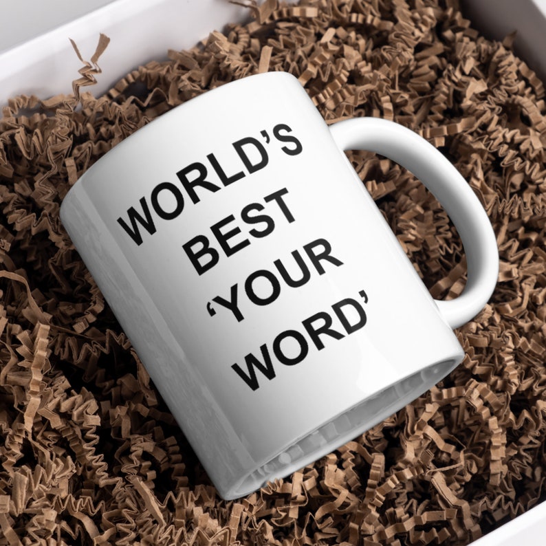 May include: White ceramic mug with black text that reads "WORLD'S BEST 'YOUR WORD'". The mug is sitting in brown packing peanuts.