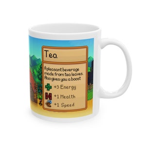May include: White ceramic mug featuring a colourful image of a video game item called "Tea". The image depicts a green background with a brown wooden sign that reads "Tea" in yellow text. The sign includes a description of the item, stating "A pleasant beverage made from tea leaves. Also gives you a boost." Below the description are three icons: a green plus sign with the text "3 Energy", a red heart with the text "1 Health", and a brown shoe with the text "1 Speed".