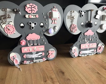Puede incluir: Dos tableros de actividades de madera gris con forma de elefante con detalles rosas y blancos. Cada tablero presenta varios elementos interactivos, como un reloj, una luz, un spinner y una cremallera. Diseñado para el juego educativo de los niños.