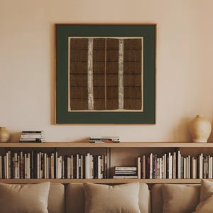 Vintage square textile wall art with intricate brown geometric patterns and cream vertical bands, surrounded by a dark green green border and hanging above a neutral sofa and built-in bookshelf in a warm modern living room.