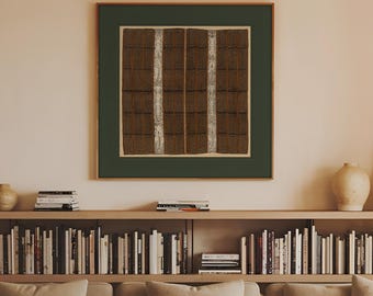 May include: A square framed wall art piece featuring a dark brown grid pattern with two vertical white textured stripes, centered above a beige sofa with three cushions, a wooden coffee table with books and bowls, and a long bookshelf filled with books.