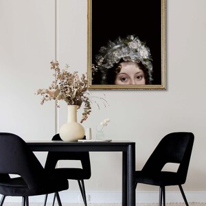 Modern dining room with a black tbale and three chairs, on the wall hangs an antique golden frame with a portraitprint showing a woman from the Regency era. Her face is at the bottom of the poster, her head can be seen from the nose up.