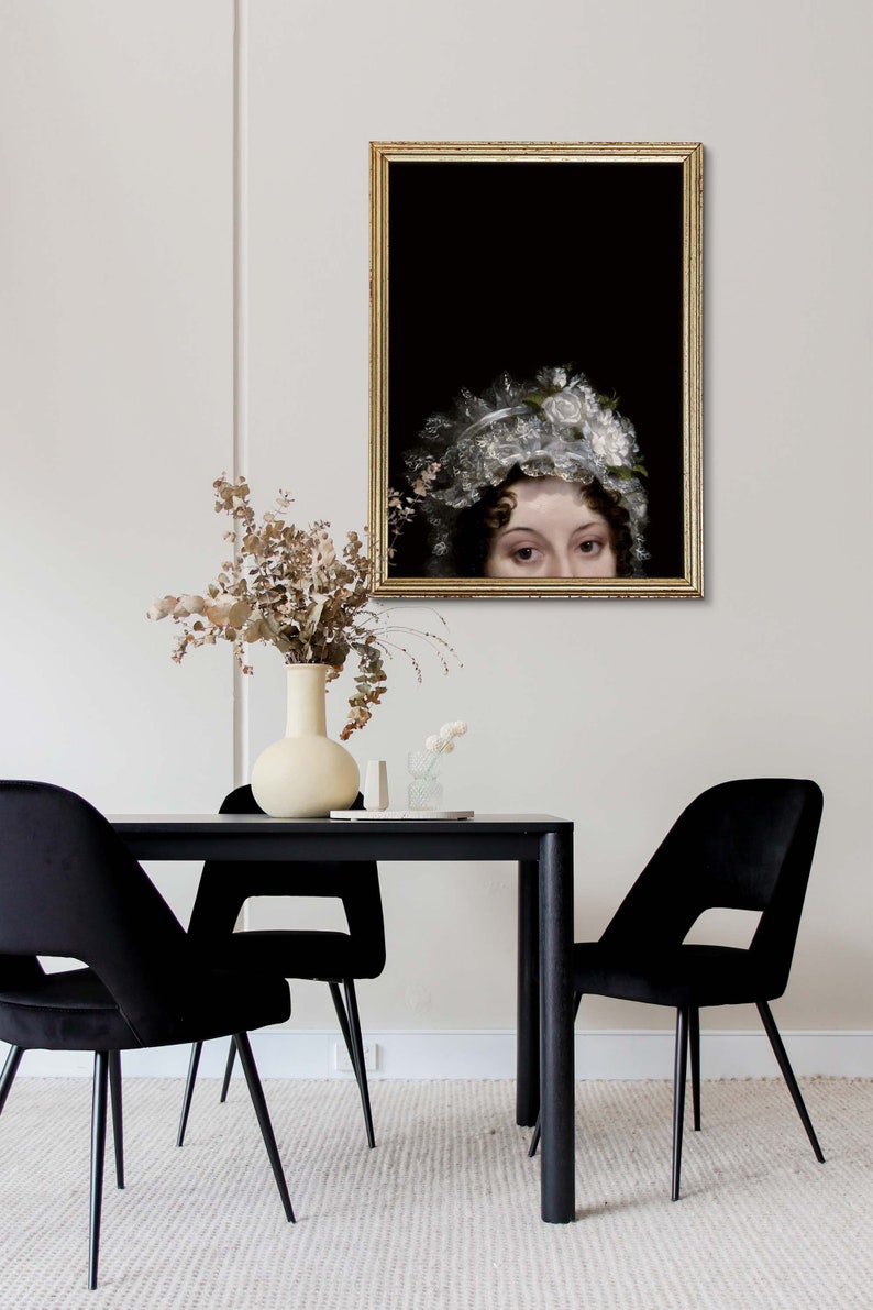 Modern dining room with a black tbale and three chairs, on the wall hangs an antique golden frame with a portraitprint showing a woman from the Regency era. Her face is at the bottom of the poster, her head can be seen from the nose up.