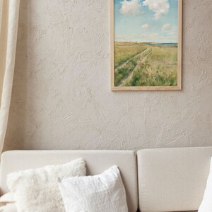 Living room with a white sofa and some ccushions on it. On the beige textured wall hangs aframe with a soft landscape of a meadow with a path through it, a seashore in the background and fluffy clouds in the blue sky.