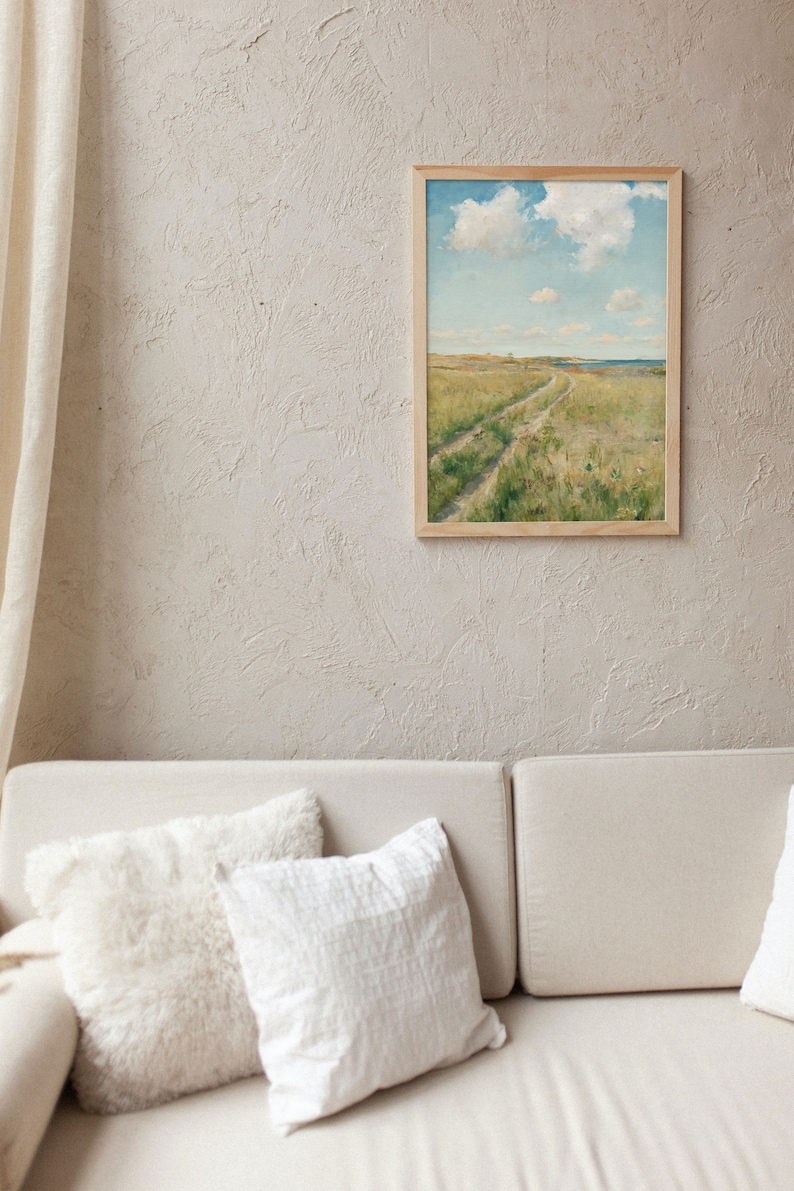 Living room with a white sofa and some ccushions on it. On the beige textured wall hangs aframe with a soft landscape of a meadow with a path through it, a seashore in the background and fluffy clouds in the blue sky.