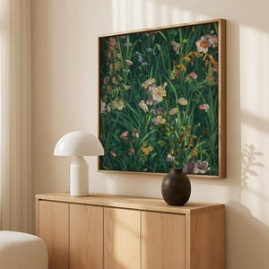 Square floral painting with green foliage and pink, yellow, and purple wildflowers, framed in wood and displayed above a light oak sideboard with a white lamp and ceramic vase.