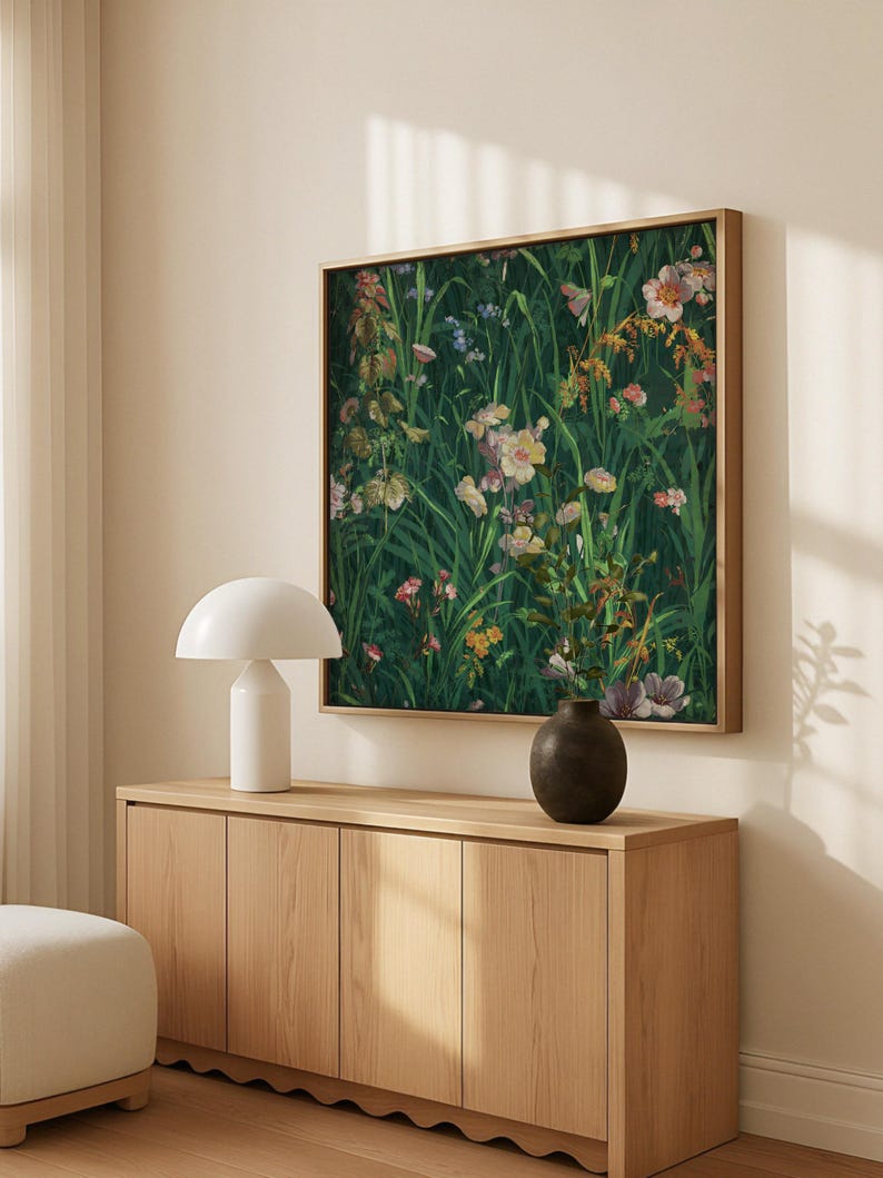 Square floral painting with green foliage and pink, yellow, and purple wildflowers, framed in wood and displayed above a light oak sideboard with a white lamp and ceramic vase.