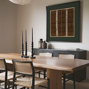 Framed square textile art print with intricate brown, black and cream geometric bands hangs above a wooden dining table. The antique woven pattern and deep green mat bring warm, earthy character to the neutral modern dining room.