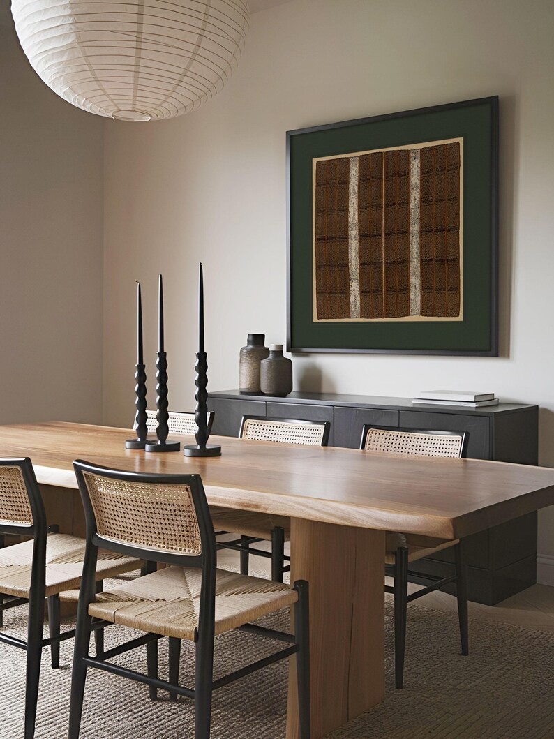 Framed square textile art print with intricate brown, black and cream geometric bands hangs above a wooden dining table. The antique woven pattern and deep green mat bring warm, earthy character to the neutral modern dining room.
