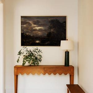 Framed moody landscape wall art with glowing moon over dark clouds and trees, styled above a wooden console table with a lamp and potted plant in a hallway.