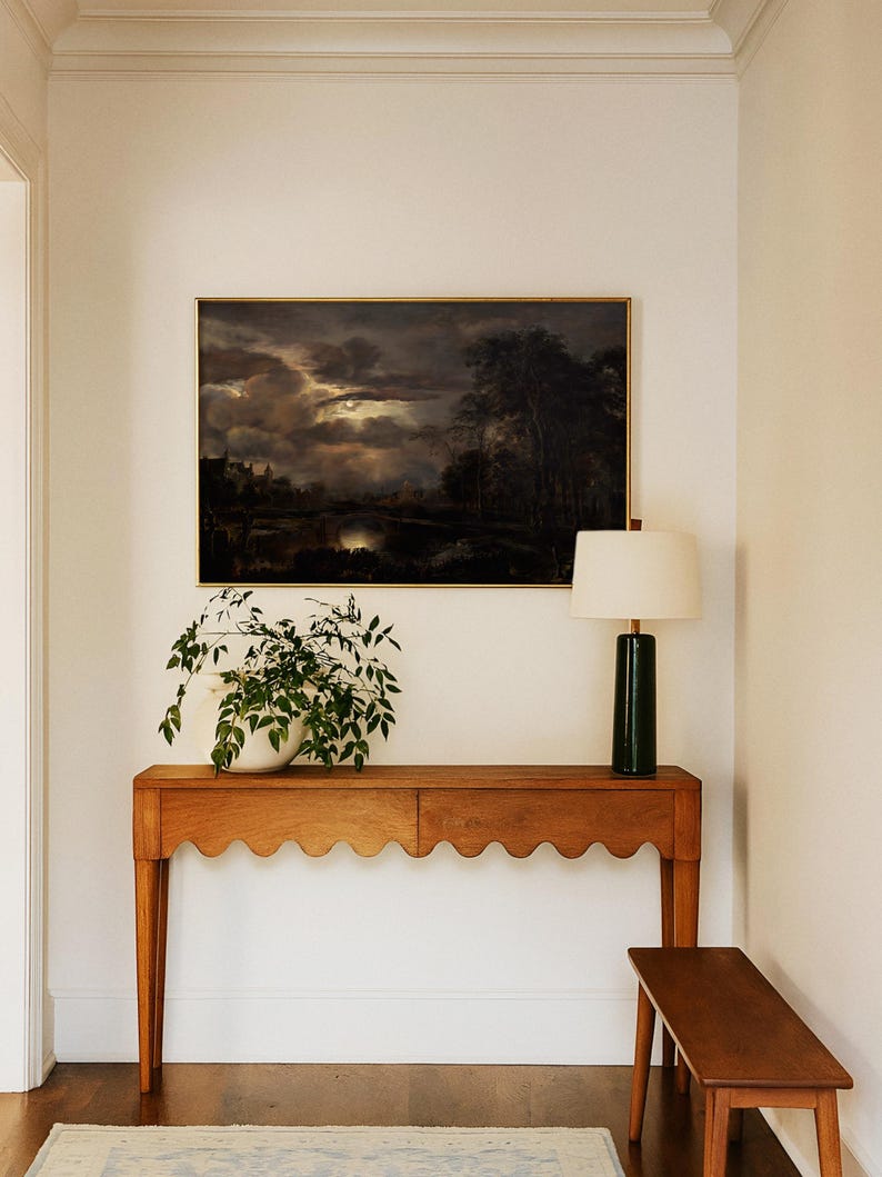 Framed moody landscape wall art with glowing moon over dark clouds and trees, styled above a wooden console table with a lamp and potted plant in a hallway.