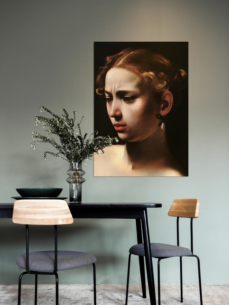 May include: A black and white portrait of a woman with a pearl earring hanging from her ear. The portrait is framed and hanging on a green wall in a dining room setting. A black table with two chairs is in the foreground.