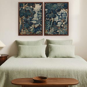 A pair of vintage verdure tapestry prints in warm wood frames hangs above a bed dressed in sage green linens. The blue botanical artwork adds calm, historic charm and creates an elegant focal point in the bedroom wall art arrangement.