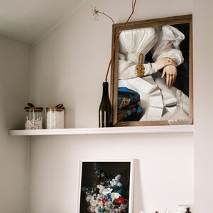 A wall niche with a shelf and a dresser with various utensils and two picture frames on it showing a dark flower still life and an altered art print of a victorian woman.