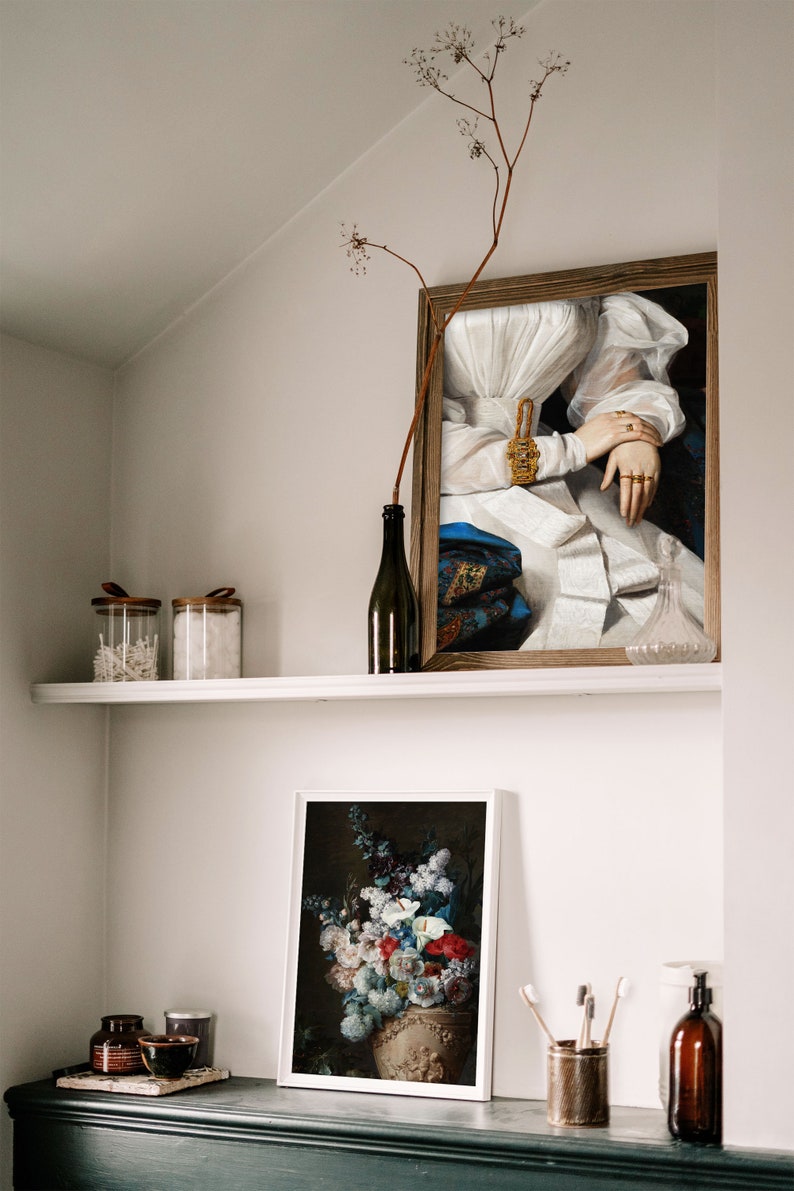 A wall niche with a shelf and a dresser with various utensils and two picture frames on it showing a dark flower still life and an altered art print of a victorian woman.