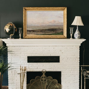 May include: A white brick fireplace with a gold metal fire screen and a set of brass fireplace tools. A framed landscape painting hangs above the fireplace.