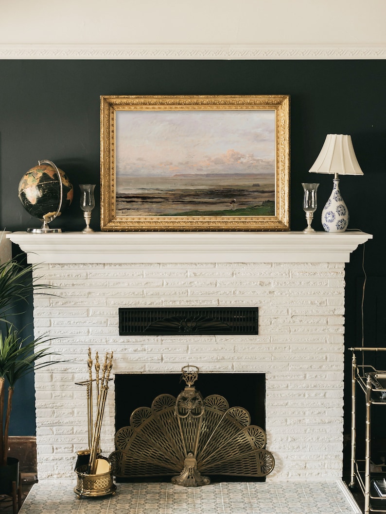 May include: A white brick fireplace with a gold metal fire screen and a set of brass fireplace tools. A framed landscape painting hangs above the fireplace.