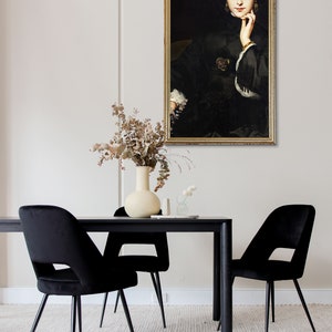 May include: A black framed painting of a woman in a black dress with a gold bracelet on her wrist. The painting is hanging on a white wall above a black dining table with two black velvet chairs.