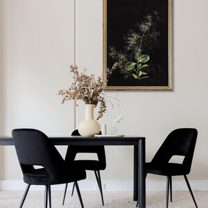 May include: A black dining table with four black velvet chairs. A vase with dried flowers sits on the table, and a framed botanical print hangs on the wall behind the table.