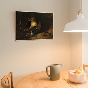 A vintage brass teapot art print hangs on a light cream-colored kitchen wall above a round wooden table. The table features a green pitcher and a ceramic bowl with fresh fruit, while a white pendant lamp softly illuminates the space.