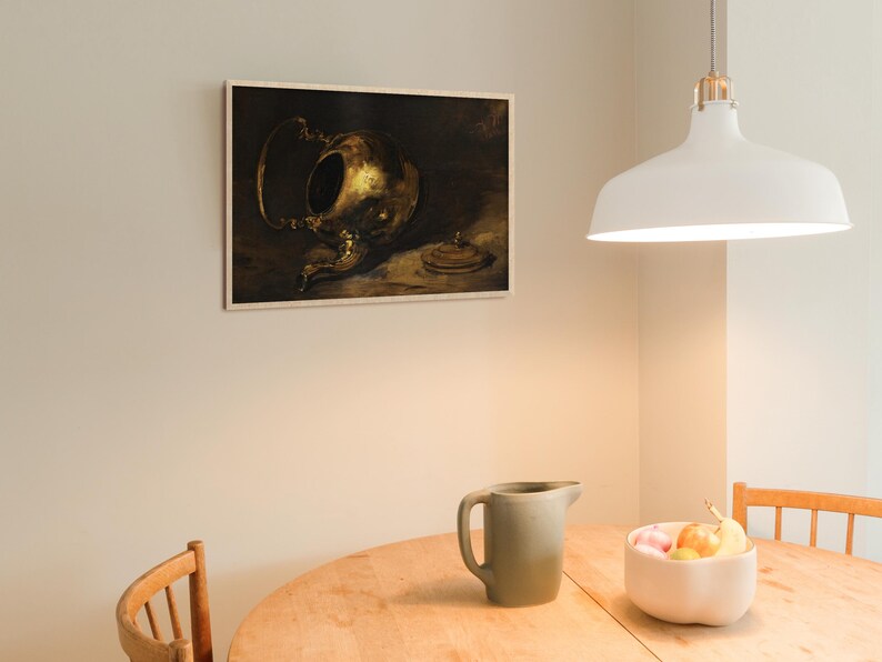 A vintage brass teapot art print hangs on a light cream-colored kitchen wall above a round wooden table. The table features a green pitcher and a ceramic bowl with fresh fruit, while a white pendant lamp softly illuminates the space.