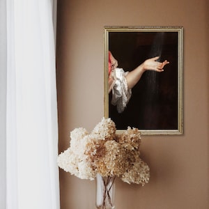 Surreal art print in a golden frame on a brown wall, in front of which there is a table with books and a vase with dried white hydrangeas