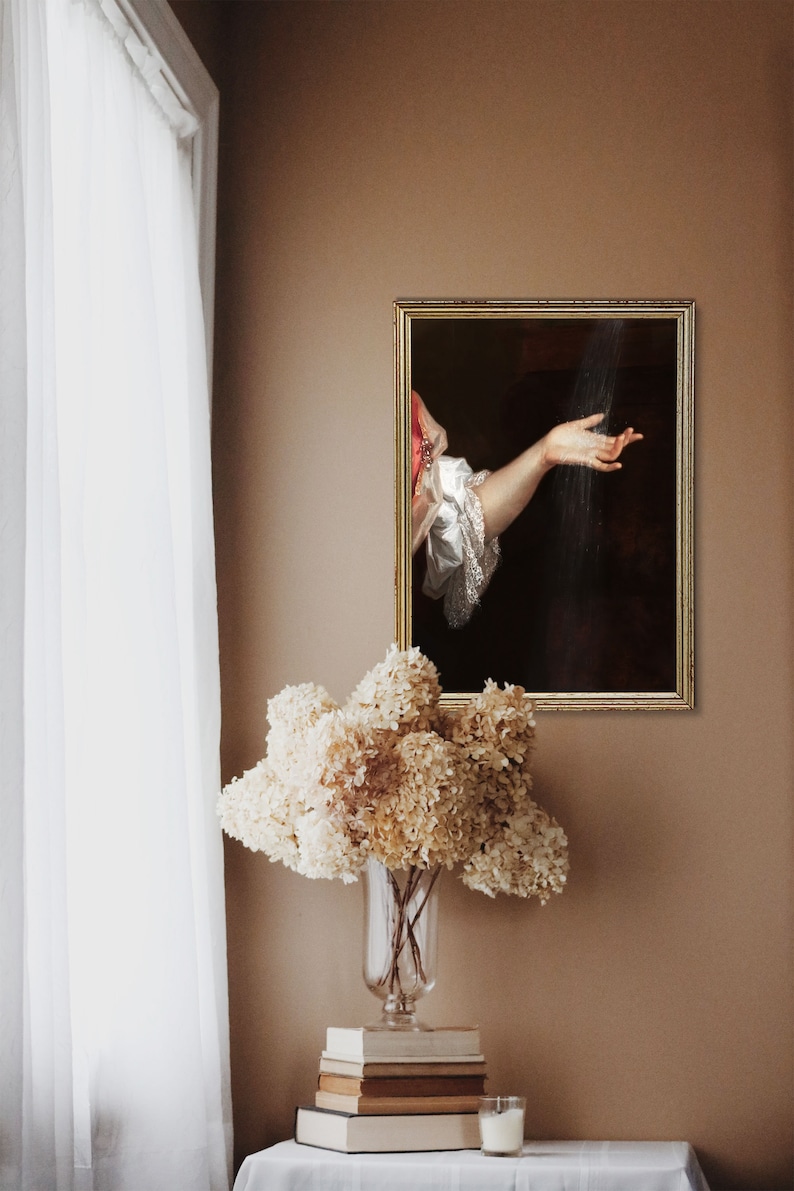 Surreal art print in a golden frame on a brown wall, in front of which there is a table with books and a vase with dried white hydrangeas