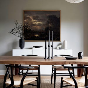 Dark academia landscape art print with moonlit clouds and a river, styled above a modern wooden dining table with minimalist chairs and black candle holders.
