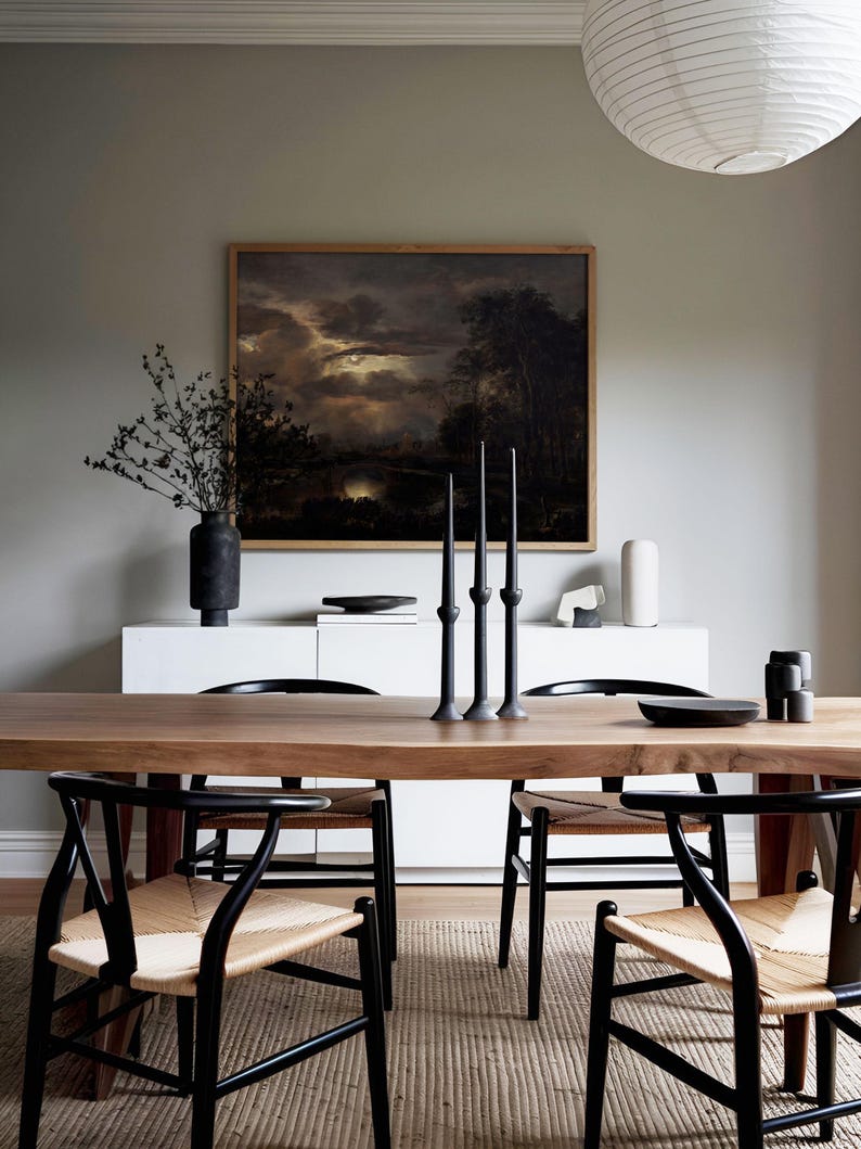 Dark academia landscape art print with moonlit clouds and a river, styled above a modern wooden dining table with minimalist chairs and black candle holders.