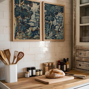 Two blue and brown verdure tapestry prints in natural wood frames hang above a rustic kitchen counter with bread and spices. The botanical textile artwork brings warmth and antique charm to the cottage-style kitchen decor.