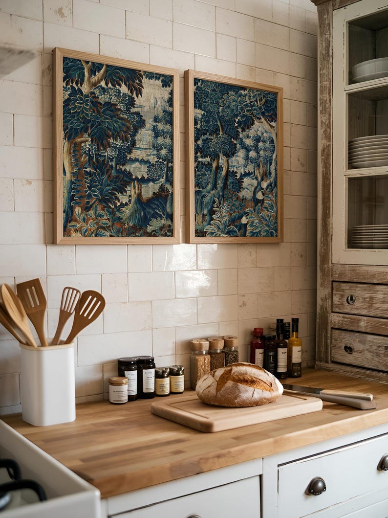 Two blue and brown verdure tapestry prints in natural wood frames hang above a rustic kitchen counter with bread and spices. The botanical textile artwork brings warmth and antique charm to the cottage-style kitchen decor.
