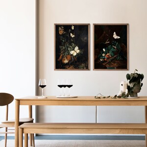 Puede incluir: Dos pinturas enmarcadas de bodegones con mariposas y fruta. Las pinturas est&aacute;n colgadas en una pared blanca sobre una mesa y un banco de madera.