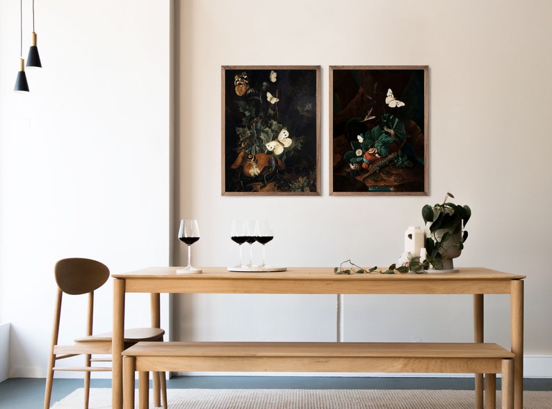 Puede incluir: Dos pinturas enmarcadas de bodegones con mariposas y fruta. Las pinturas est&aacute;n colgadas en una pared blanca sobre una mesa y un banco de madera.